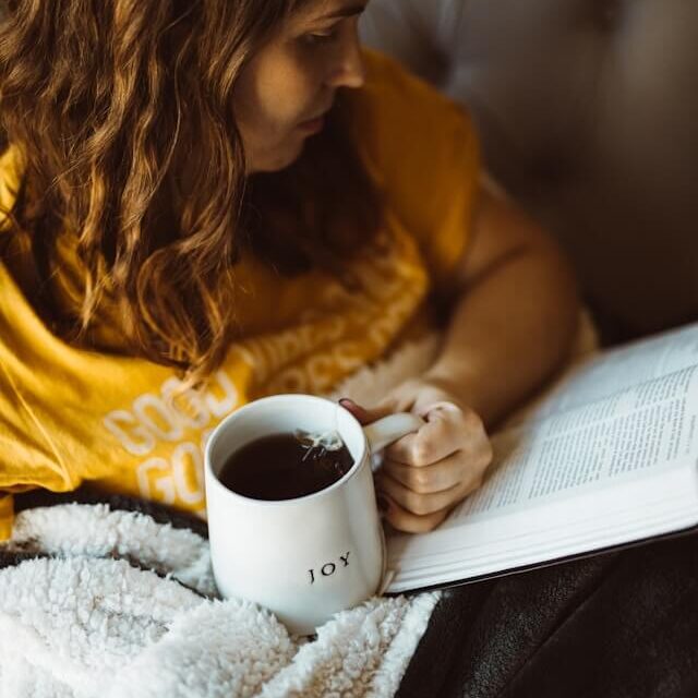 A girl reading a book while holding a coffee mug that says "joy" on the front of it (Photo Credit: Nathan Dumlao)