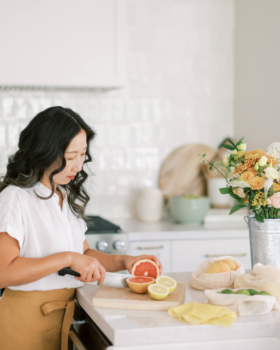 A woman slicing grapefruit and lemons on a wooden board in a modern kitchen, surrounded by reusable produce bags and a bouquet of flowers. (Photo Credit: Haute Stock)