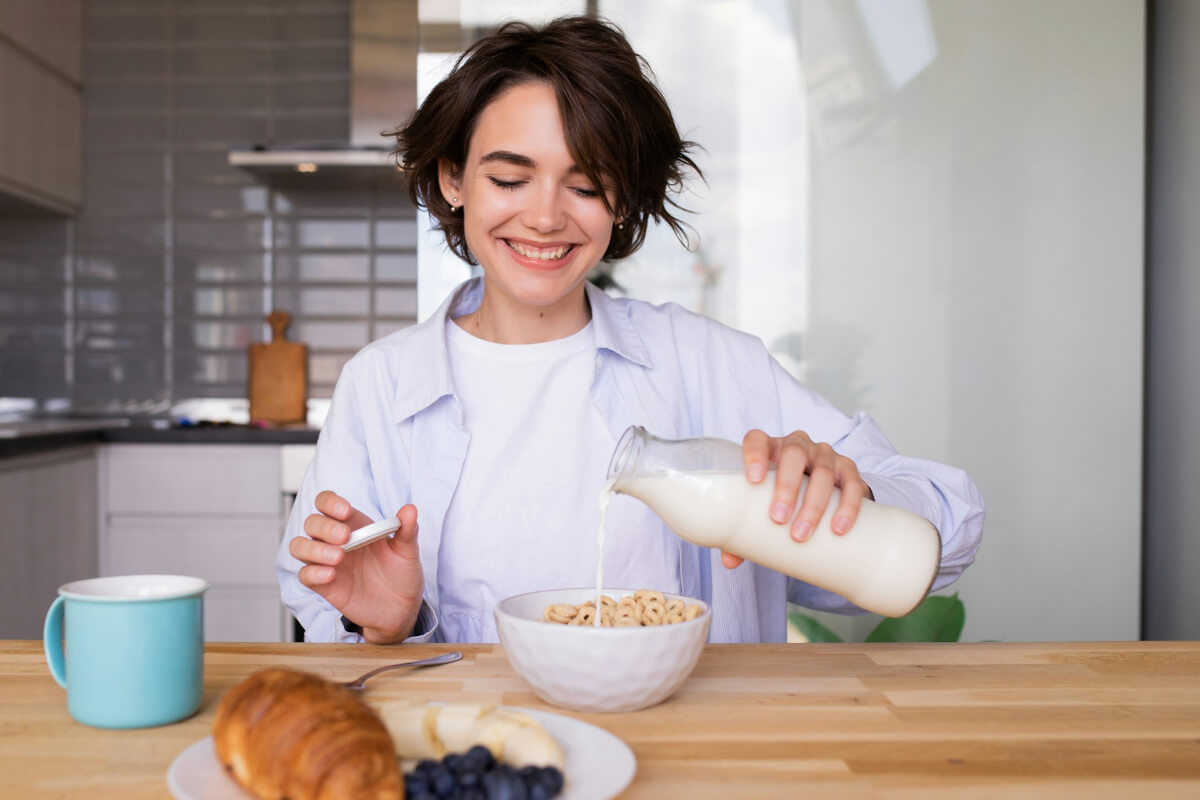 Smiling young woman pouring milk from a glass bottle over cereal in a white bowl at a wooden table. (Photo Credit: Look Studio)