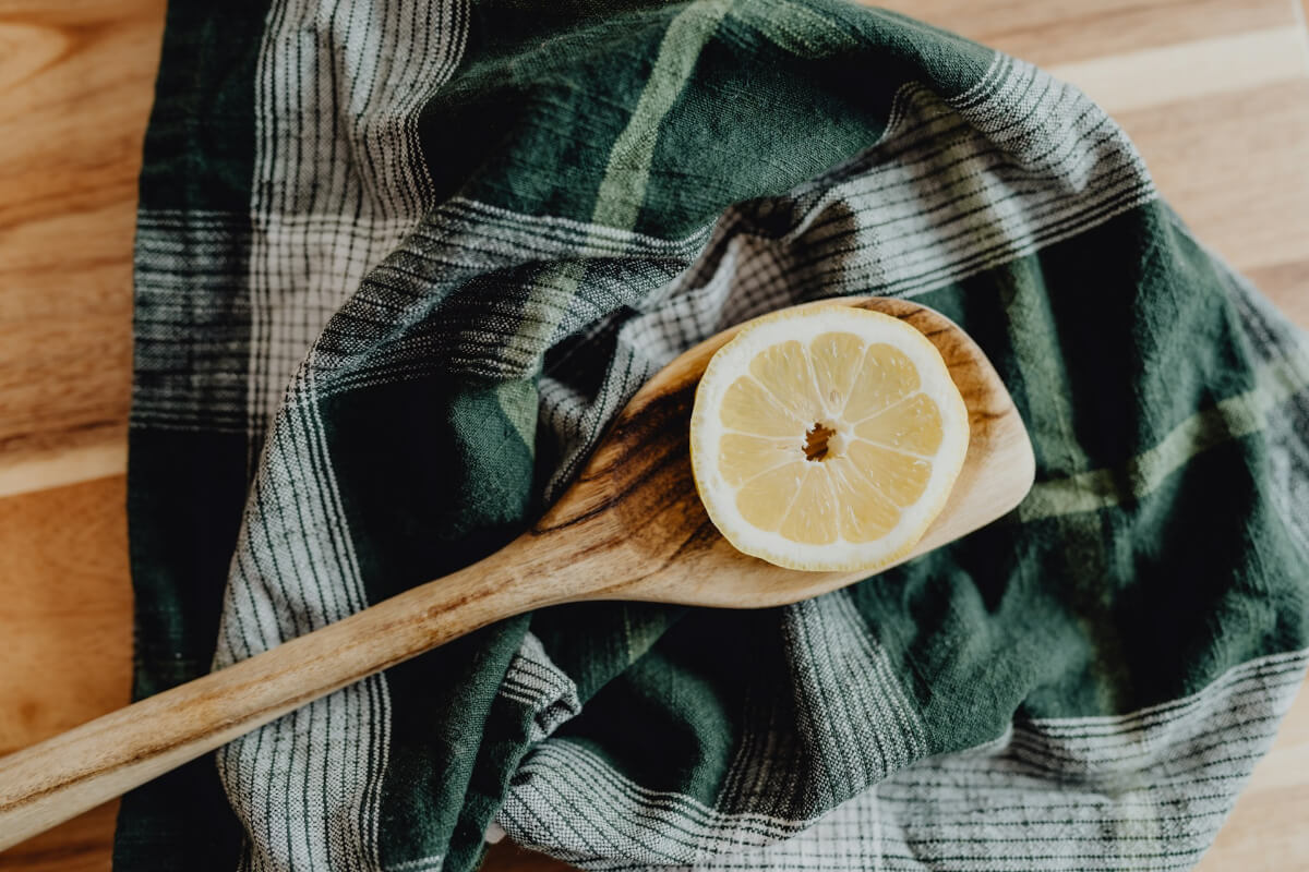 A rustic wooden spoon resting on a dark green and white plaid linen cloth draped over a butcher block, holding half a fresh lemon. (Photo Credit: Kelly Sikkema)