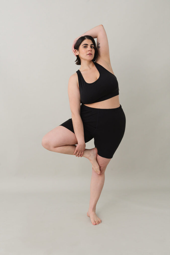 A woman poses in a black matching bra and bike shorts set by ODDOBODY, made with certified fair-trade cotton. She stands barefoot in a tree pose against a light neutral background, with one arm raised above her head and a floral tattoo visible on her upper arm. The scoop-neck bra and high-rise shorts showcase the brand’s ethical and minimalist approach to comfort-focused basics. (Photo Credit: ODDOBODY)