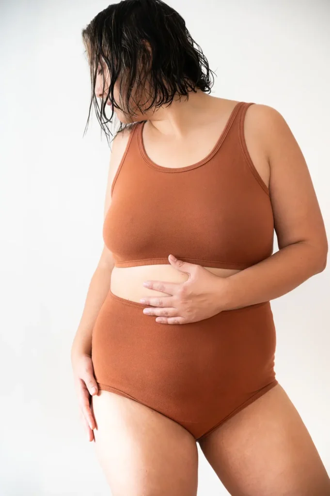 A woman with wet hair wears the rust-colored Coco Bra and matching high-rise briefs by Archer Wren, made from 92% organic cotton and 8% spandex. The set is soft, flexible, and size-inclusive, shown here in a minimalist studio setting. Ethically handmade in women-owned U.S. factories and dyed with AZO-free dyes, the design balances comfort, support, and inclusive sustainability. (Photo Credit: Archer Wren)