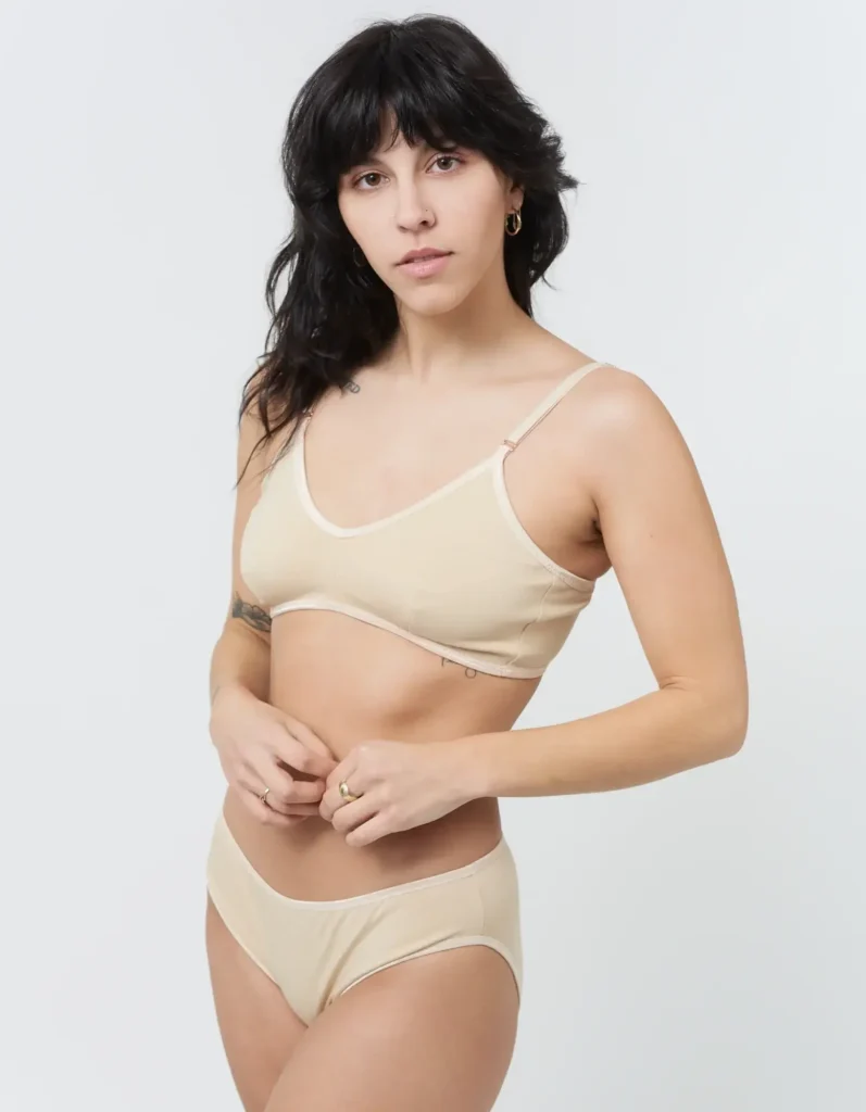 A woman wears a nude triangle bra and matching briefs by Brook There, made from 94% GOTS-certified organic cotton and 6% spandex. The minimalist set features soft seams, subtle contrast trim, and a breathable, U.S.-made construction that reflects the brand’s small-batch, sustainable ethos. (Photo Credit: Brook There)