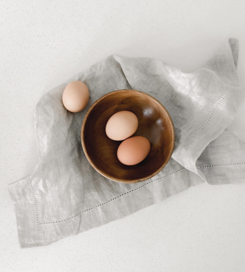 A minimalist top-down image of three brown eggs placed in a wooden bowl, with a fourth egg resting on a soft grey linen cloth over a white countertop. The neutral tones and soft textures create a calm, airy aesthetic (Photo Credit: Haute Stock).