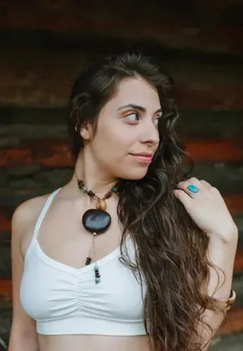 A woman with long wavy hair wears a white cami-style bra by Circle Creations, made from a breathable blend of hemp, organic cotton, and Lycra. She poses outdoors in front of a rustic wooden backdrop, accessorized with a bold natural pendant and turquoise ring. The bra features gentle ruching at the center and thin straps, reflecting the brand’s handmade, small-batch ethos from Oregon. (Photo Credit: Circle Creations)