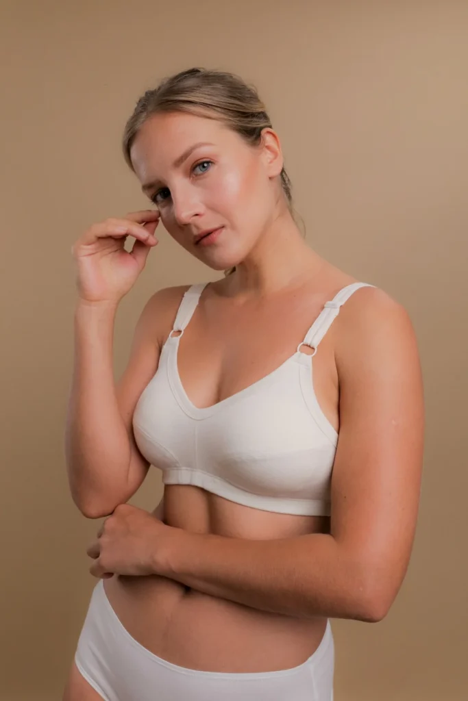 A woman wears a white wire-free bra and matching briefs made from 100% organic cotton by Cottonique. Designed for sensitive skin, the bra features wide adjustable straps with plastic ring connectors, soft seaming for structure, and no latex or spandex. She poses against a warm beige background, highlighting the bra’s gentle, non-restrictive fit and natural comfort. (Photo Credit: Cottonique)