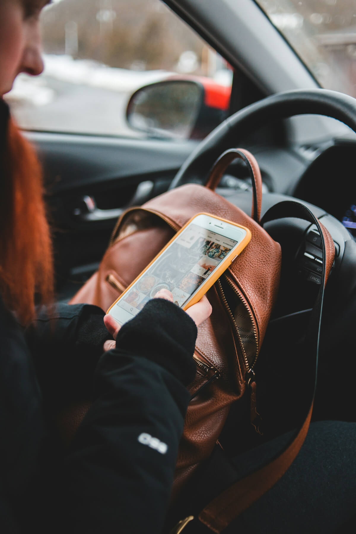 Close-up of a person using a smartphone while seated in a car, resting the phone on a brown leather backpack placed on their lap. The phone screen shows a social media feed with various photos, and the person is dressed in a black jacket. (Photo Credit: Erik Mclean)