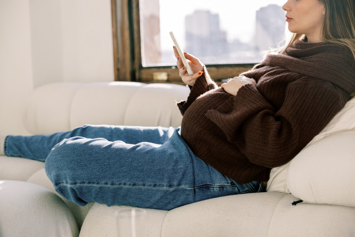A pregnant woman lounges on a white cushioned sofa, dressed in a cozy brown knit sweater and blue jeans. She holds a smartphone and looks relaxed, with city buildings visible through the nearby window. (Photo Credit: Haute Stock)