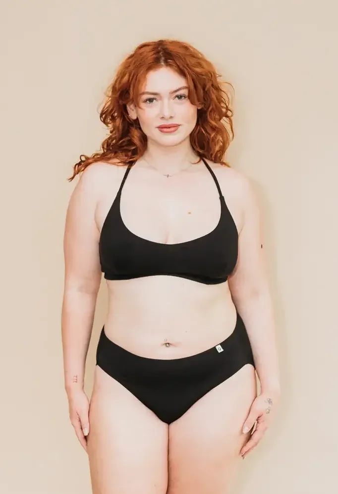 Woman with curly red hair models a black criss-cross bralette and matching briefs while standing against a beige studio backdrop. (Photo Credit: Q For Quinn)
