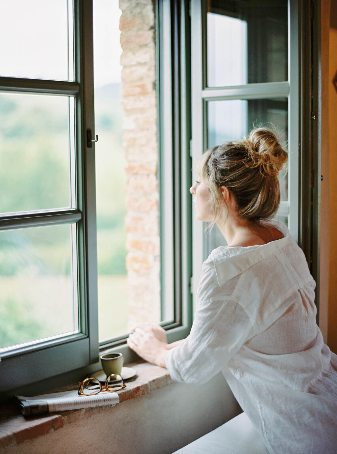 Woman in a white linen blouse gazes out of an open window toward a green landscape, with a coffee cup, glasses, and magazine resting on the brick window ledge. (Photo Credit: Haute Stock)