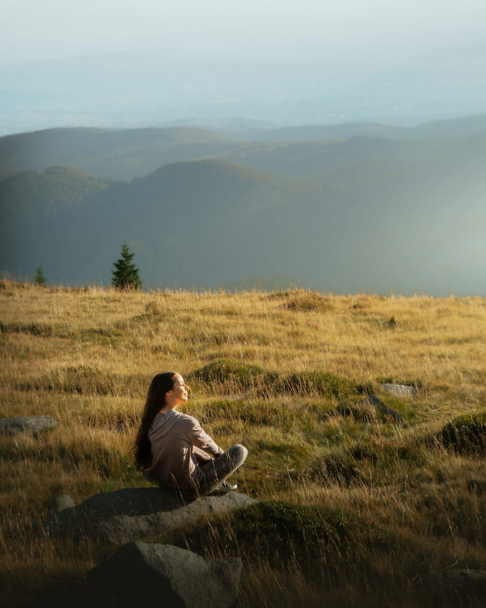 Woman seated on rocky grassy hillside gazing out over misty rolling mountain ridges, golden sunlight bathing the scene. (Photo Credit: Inspa Makers)