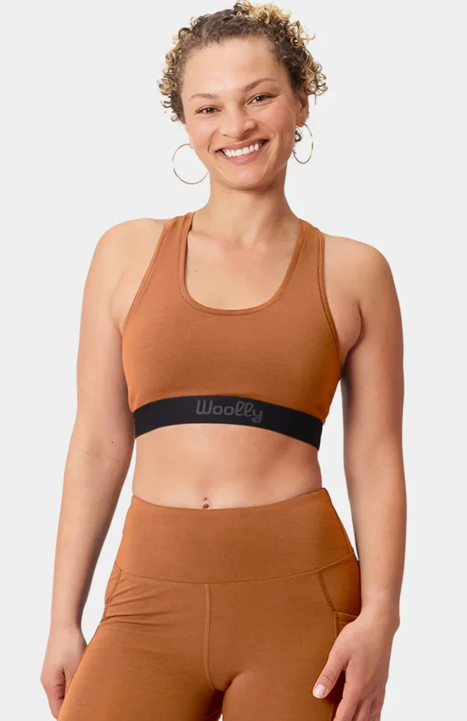 A woman smiles while wearing a rust-colored merino wool racerback sports bra by Woolly, featuring a wide black elastic band with the brand name printed across it. Made from 95% RWS-certified merino wool and 5% elastane, the bra offers natural moisture-wicking, odor resistance, and breathable support for everyday or active wear. (Photo Credit: Woolly)
