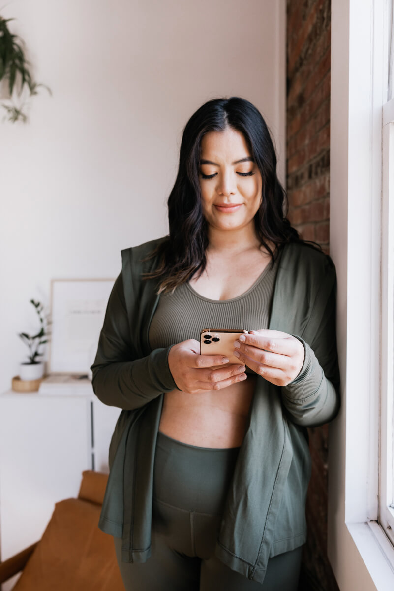 An Asian woman with long dark hair looks down at a rose gold smartphone held in both hands. She wears a coordinated olive green ensemble consisting of a cardigan over a ribbed sports bra and matching pants. Natural light streams from a nearby window, with exposed brick visible in the soft-focus background. (Photo Credit: Haute Stock)