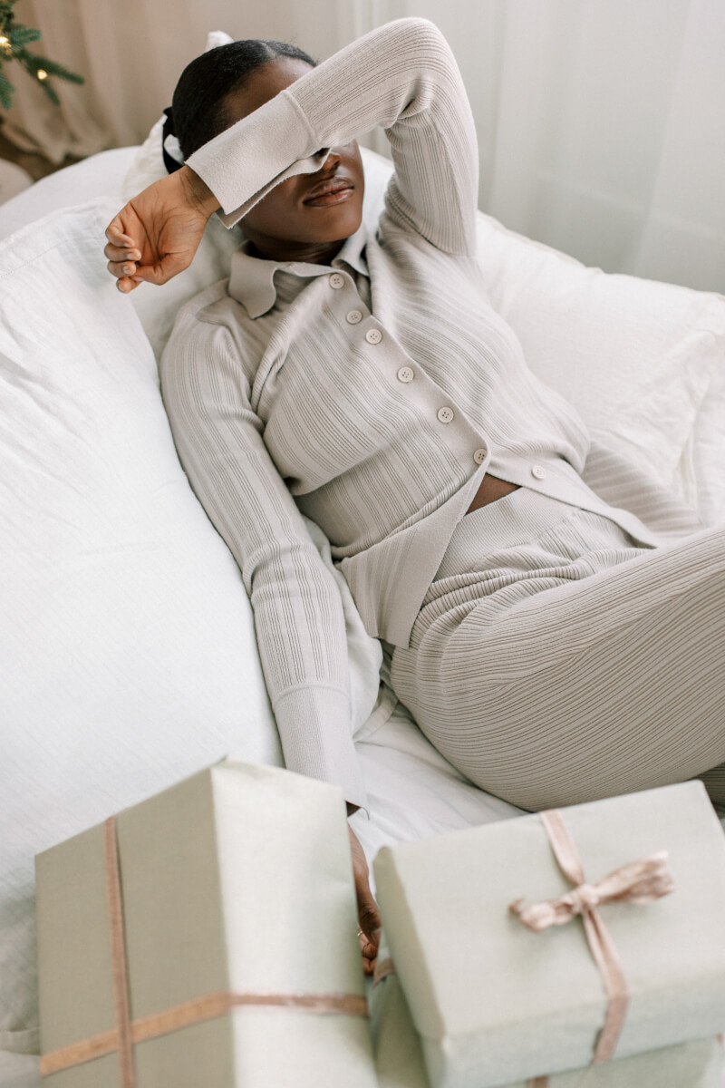 A Black woman on a white bed wearing a textured beige ribbed lounge set, covering her eyes with one arm. Several wrapped gifts in cream, sage green, and blush tones with ribbon accents are arranged in front of her. (Photo Credit: Haute Stock)