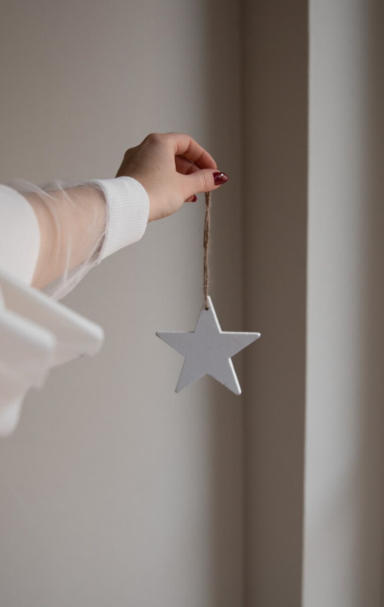A hand in a white knit sleeve with dark red nail polish holds a simple white wooden star ornament suspended from natural twine against a soft neutral wall background. (Photo Credit: Haute Stock)