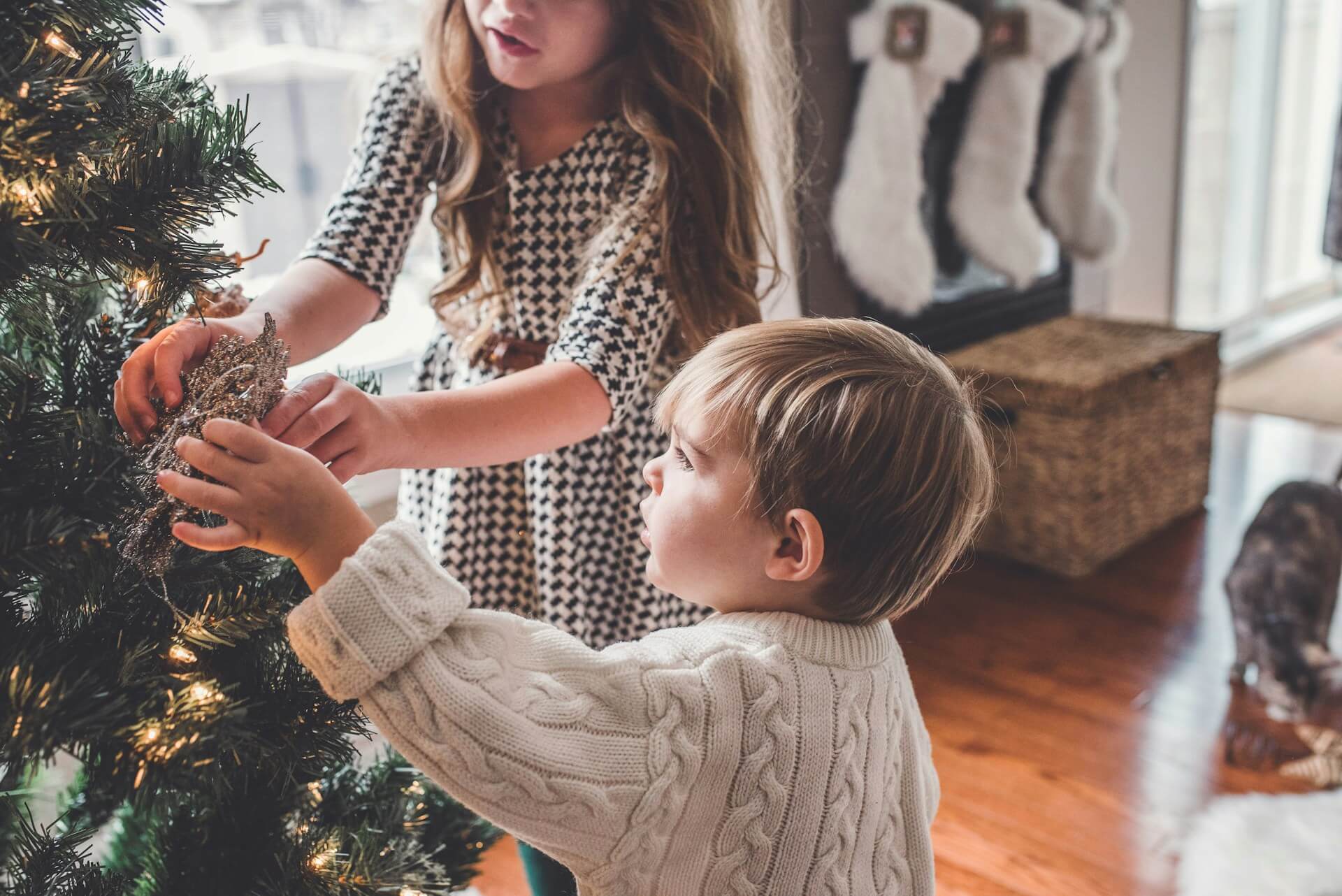 A blonde child in a cream cable-knit sweater reaches up to hang a glittery ornament on a Christmas tree adorned with white lights. Behind him, a little girl with long brown hair in a black and white houndstooth dress helps guide the decoration into place. The cozy scene unfolds on warm hardwood floors with holiday stockings softly blurred in the background. (Photo Credit: Paige Cody)