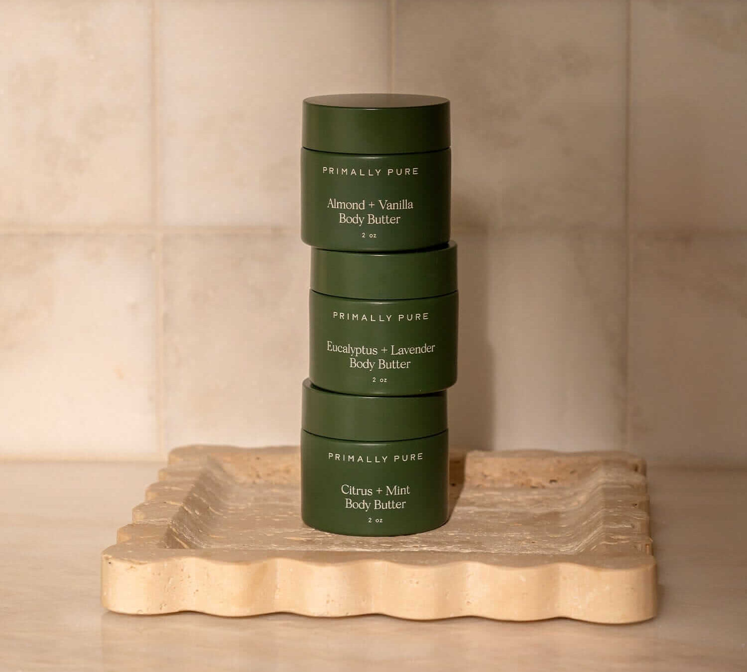 Three Primally Pure body butter jars stacked on wooden platform including almond vanilla, eucalyptus lavender, and citrus mint varieties in dark green 4oz containers (Photo Credit: Primally Pure)