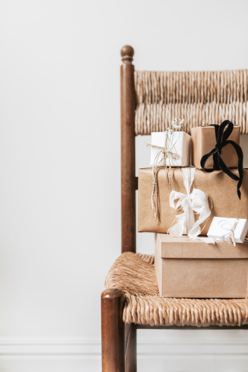A stack of wrapped gifts in kraft paper adorned with white ribbons, black bows, and sprigs of dried baby's breath sits on a natural woven rush seat chair with wooden frame against a white wall. (Photo Credit: Haute Stock)