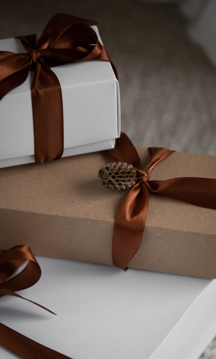 A close-up shows elegantly wrapped gifts stacked together, featuring white and kraft paper wrapping tied with rich copper-brown satin ribbons. A small natural pinecone adorns one of the packages as a rustic embellishment against the minimalist wrapping. (Photo Credit: Haute Stock)